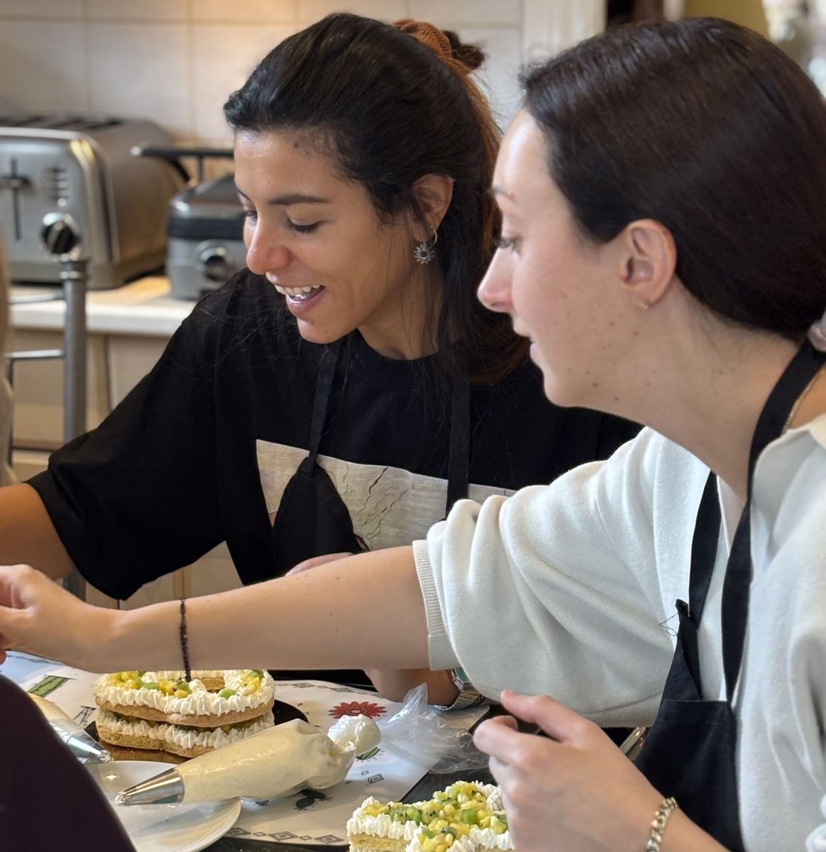 Deux amies qui garnissent leur letter cake à la poche à douille pendant un atelier de pâtisserie à Montréal